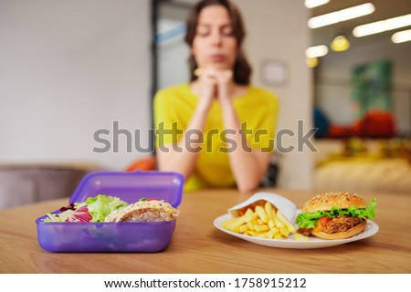 Vegetables or burger. Young woman sitting at table indoors choosing between fresh chopped vegetables in lunchbox or burger on plate