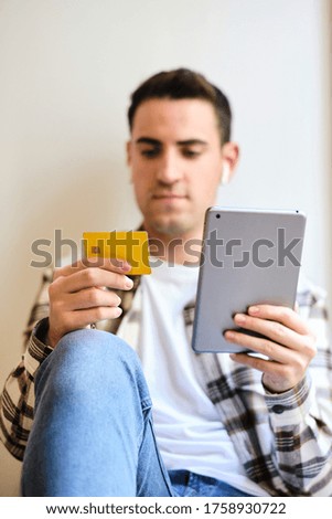 Young man shopping and paying online on digital tablet with credit card while relaxing at home listening music with earphones.