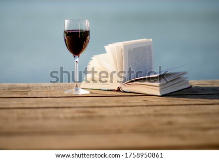 A glass of red wine and a book on the wooden pier in front of blue water background. 
