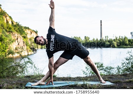 Inspired man doing yoga asanas on quarry lake. Young citizen exercising outside and standing in yoga side angle pose. Fitness outdoors and life balance concept.