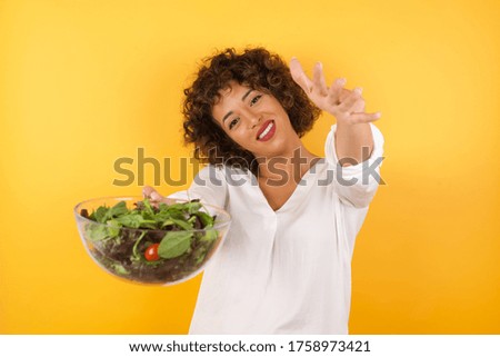 Young arab woman with curly hair wearing shirt holding a salad standing over isolated yellow background looking at the camera smiling with open arms for hug. Cheerful expression embracing happiness.