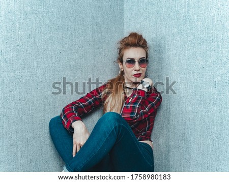 A beautiful girl in a red shirt with glasses and with a clock on her hand sits near the wall with gray wallpaper