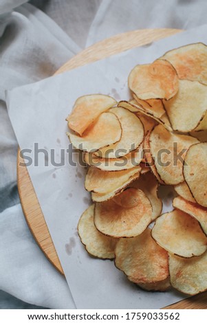Fresh homemade deep fried crispy potato chips in white box on a wooden tray, top view. Salty crisps scattered on a table for a tasty snack break and party movie time.