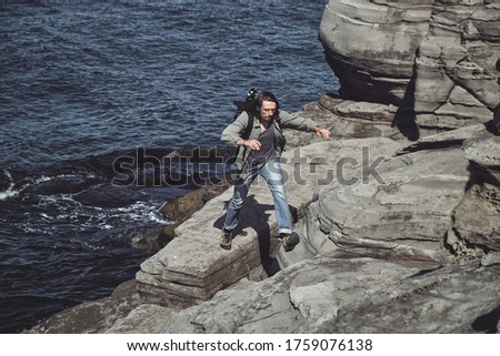 young long-haired man with a backpack and a photo tripod jumps over an abyss in a rock on the seashore. Travel and outdoor concept.