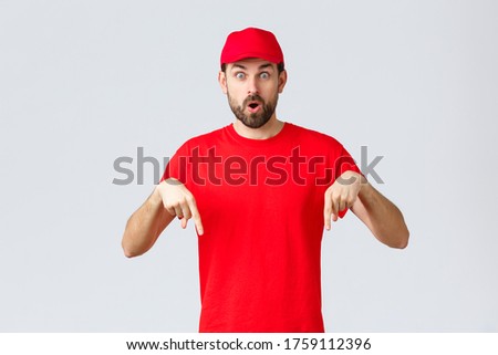Online shopping, delivery during quarantine and takeaway concept. Surprised and intrigued courier inform clients, employee in red uniform cap and t-shirt pointing fingers down, say wow