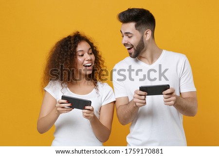 Funny young couple two friends european guy african american girl in white t-shirts posing isolated on yellow background. People lifestyle concept. Mock up copy space. Play game with mobile phone