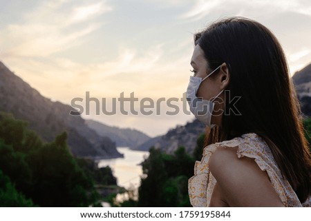 Beautiful woman wearing a mask looking at a reassuring landscape at sunset while on rural vacation in coronavirus time, far from the city. Selective focus