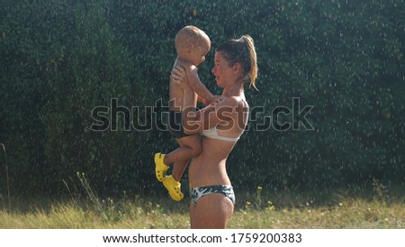 Young mother standing under water spray and holding smiling little son in her arms. Mother and smiling child looking at each other