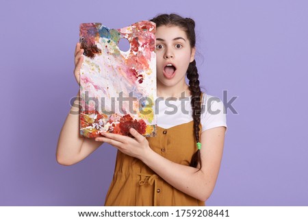 Indoor shot of creative young woman painting in her studio. Female artist in her workshop holding colors palette near her face, stands with widely open mouth, expresses shock, poses against lilac wall