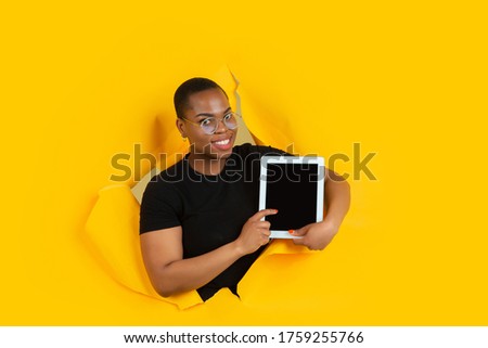 Shows blank tablet screen. Cheerful african-american young woman poses in torn yellow paper background, emotional. Breaking on, breakthrought. Concept of human emotions, facial expression, sales, ad.