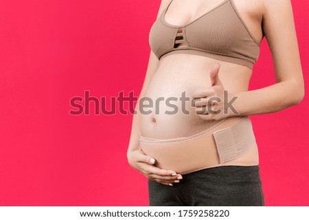 Close up of pregnant woman showing thumb up and wearing elastic pregnancy bandage to make the pain go away at pink background with copy space. Orthopedic abdominal support belt concept.