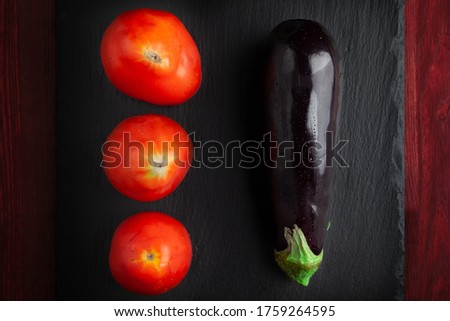 Raw eggplant with three tomatoes on black schist board on dark wooden tanle. Flat lay. Low key.