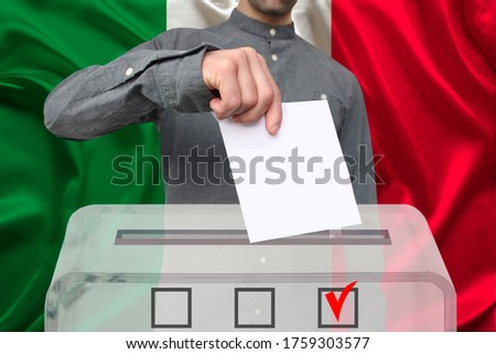 male voter lowers the ballot in a transparent ballot box on the background of the national flag of italy, concept of state elections, referendum