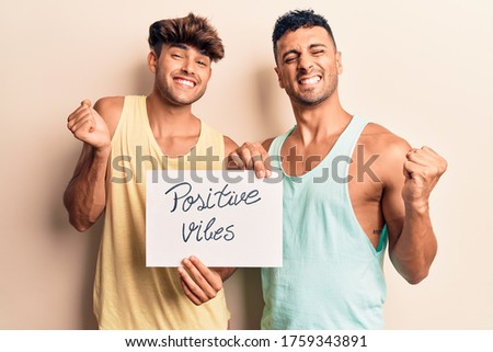 Young gay couple holding positive vibes banner screaming proud, celebrating victory and success very excited with raised arm 