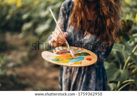 Close up hands of female artist holding brush and palette with oil paints. Blurred background with easel in sunflower field.