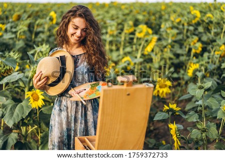 A young woman with curly hair and wearing a hat is painting in nature. A woman stands in a sunflower field on a beautiful day.
