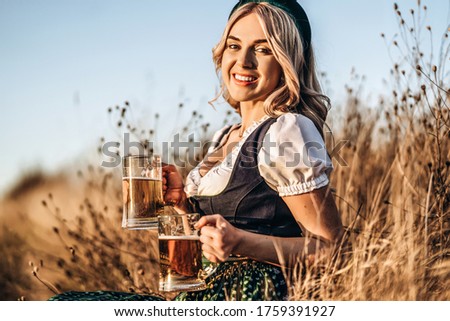 Pretty happy blonde in dirndl, traditional festival dress, holding two mugs of beer outdoors in the field with blurred background. Oktoberfest, St. Patrick’s day, international beer day concept.