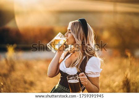 Pretty happy blonde in dirndl, traditional festival dress, holding two mugs of beer outdoors in the field with blurred background. Oktoberfest, St. Patrick’s day, international beer day concept.