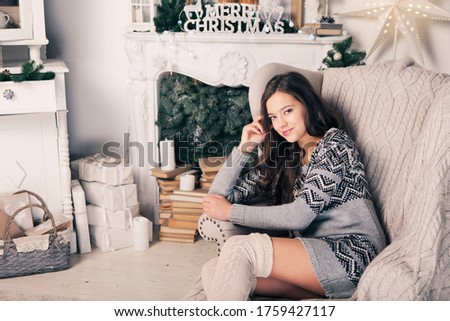 Beautiful girl near fireplace with Christmas tree, candles sits in comfortable armchair. Scandinavian cozy home decor