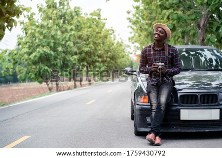 African man traveler is holding camera and standing near car freelancing on the road