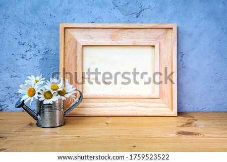 wooden picture frame on table with miniature watering can and daisies on blue background