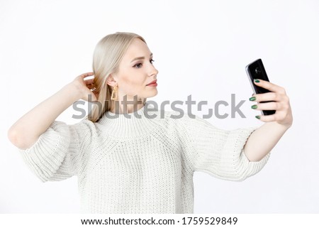 young blonde girl with a slight smile takes a selfie on a white background