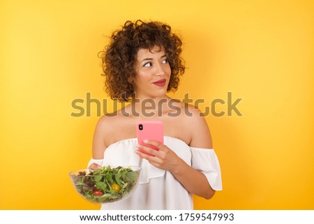 Image of a happy young beautiful woman holding a salad posing isolated over bright background listening to music with earphones using mobile phone.