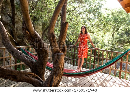 A woman in a red dress stands on the terrace of a wooden house and enjoys the view, Veranda in the shade of trees, Balcony with garden view.