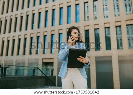 Attractive business woman in a blue jacket with a black folder in her hands smiling and makes a phone call against the background of office buildings