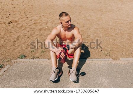 Young bearded man sitting on basketball court outdoors with ball holding bottle drinking electrolytes drink looking camera smiling happy