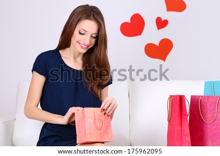 Beautiful young woman sitting on sofa with shopping bags on gray background