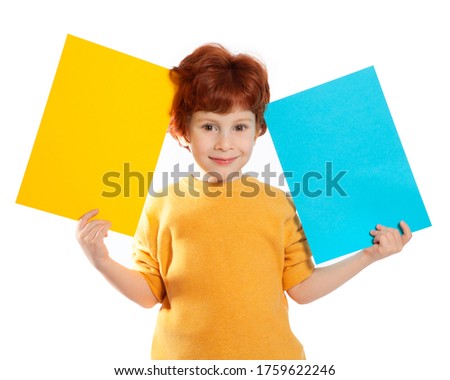 A beautiful European boy with red hair holds two sheets of colored paper on a white background, smiling. Space for text, banners, labels, and ads.