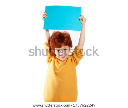 A beautiful European boy with red hair holds a blue sheet of paper above his head on a white background and smiles. Space for text, banners, labels, and ads.