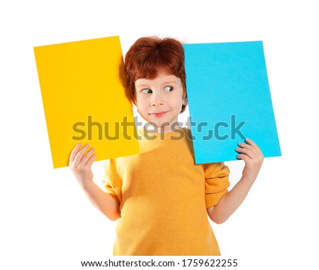 A beautiful European boy with red hair holds two sheets of colored paper on a white background, smiles, is surprised, looks away. Space for text, banners, labels, and ads.