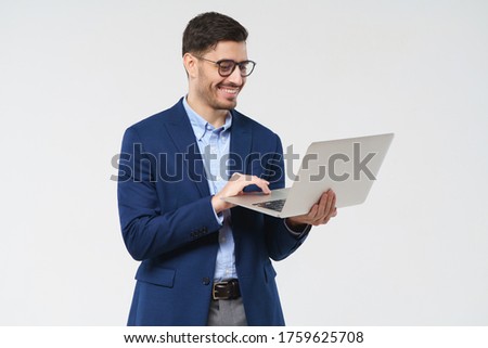 Young male teacher wearing eyeglasses, holding open laptop, looking at content on screen with happy smile, isolated on gray background