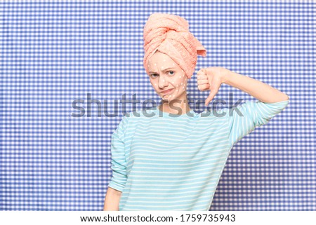 Portrait of unhappy sad girl with anti-acne skincare product on face, with towel on head, showing thumb down sign of dislike, over shower curtain background. Care for skin and hair. Beauty concept