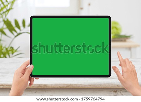 Isolated human left hand holding black tablet media device with white green screen mockup and wooden table in kitchen