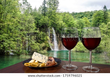 Two glasses of wine with charcuterie assortment waterfall background in Croatia. Glass of red wine with different snacks - plate with ham, sliced, blue cheese. Romantic celebration.