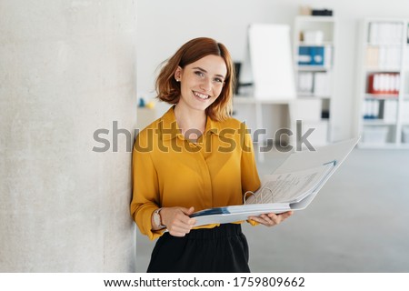 Attractive young office worker holding a large open binder as she looks at the camera with a sweet friendly smile Royalty-Free Stock Photo #1759809662