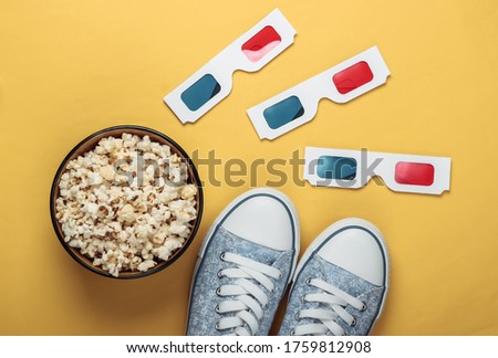 Anaglyph disposable paper 3d glasses with old school style sneakers and popcorn bowl on yellow background. Top view. Retro 80s