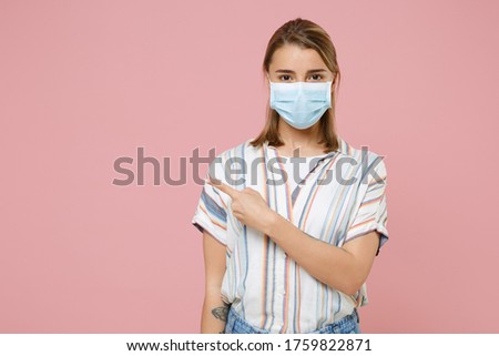 Young blonde woman girl in casual striped shirt sterile face mask isolated on pink background. Epidemic pandemic coronavirus 2019-ncov sars covid-19 flu virus concept. Pointing index finger aside