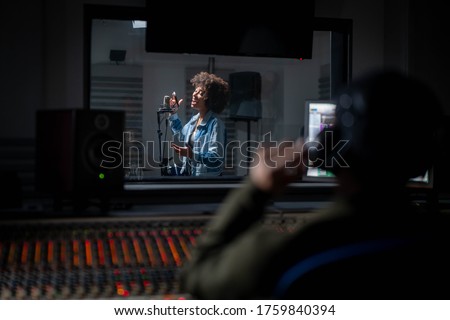 Back view of professional male sound producer with headphones is recording a new song with young african female singer in a music studio.