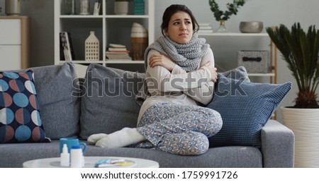 Pretty sad young ill woman in the warm scarf shivering while sitting on the couch in the living room as she having a fever. Indoors Royalty-Free Stock Photo #1759991726