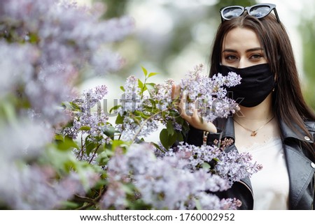 Girl in a medical mask on a background of blooming lilacs. Black mask. Protection against virus, flu. Coronavirus protection. epidemic of coronavirus. the aroma a tree in the garden Spring allergy.