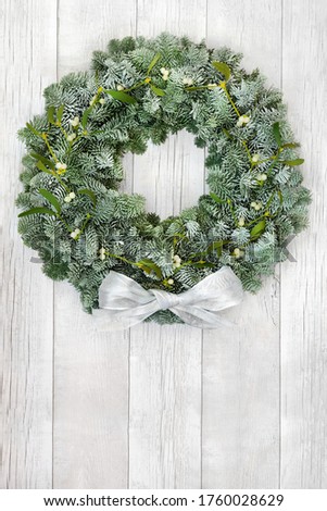 Snow covered spruce fir & mistletoe winter wreath with white bow on rustic wood front door background. Composition for solstice, Christmas & New Year. Copy space.