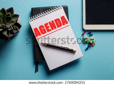 AGENDA written in red in a notebook on a blue background near a tablet and a flower in a pot