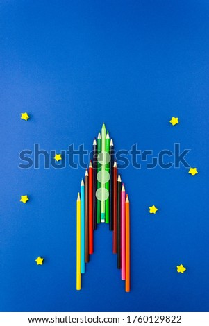 Rocket made of pencils on blue cardboard is decorated with wooden stars and paper confetti in the form of portholes. Preparing for school and buying office supplies. Vertical, copy space