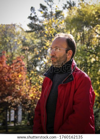 A beautiful portrait of a brutal tanned man in glasses, a guy, a brunette, with bristles and a red jacket, which stands in nature or in the city on a summer or autumn-spring day.