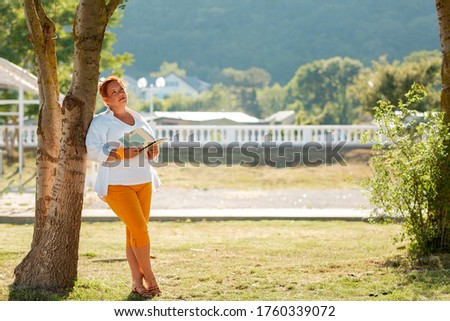 Education and leisure. A woman stands in the Park, leaning against a tree and holding a book, looking thoughtful. Copy space