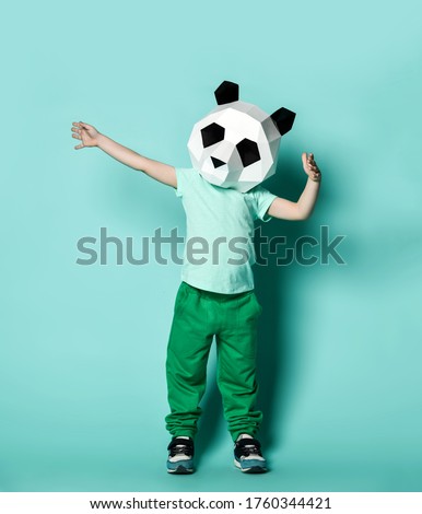Kid boy in panda mask white blank t-shirt and green pants is posing with arms spread wide holding something big on pastel blue background